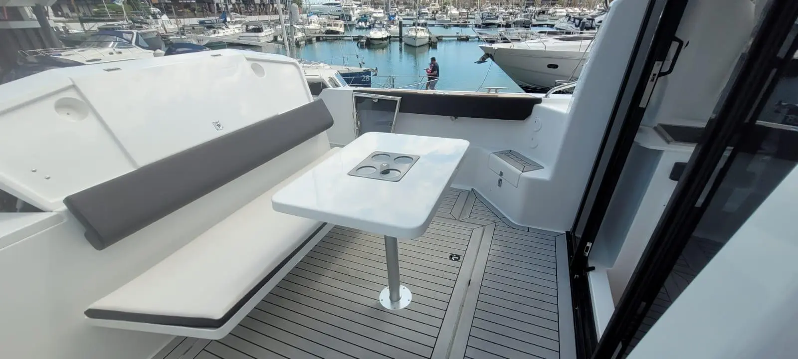 Modern boat interior with a white table, cushioned seating, and views of a marina filled with boats.